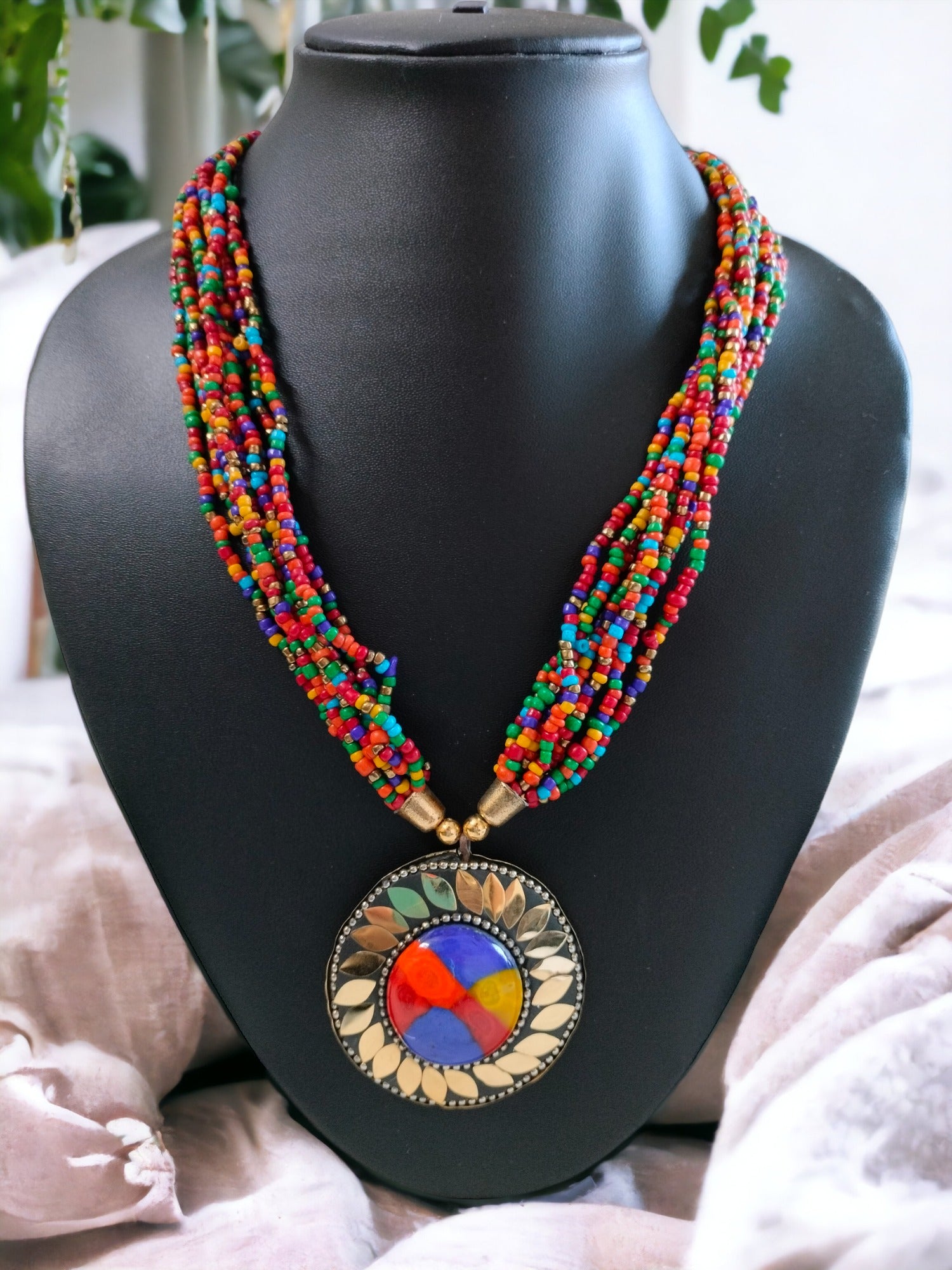Vibrant multi-colored bead necklace with a large circular pendant featuring a colorful stained glass design, showcased on a black mannequin against a soft fabric backdrop."